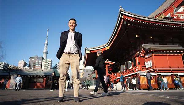 Yohay Wakabayashi standing in front of Tokyo street wtih buildings and people behind him.